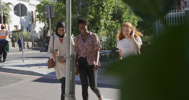 Three women from diverse backgrounds are walking and chatting happily in a sunny urban street. One is wearing a hijab, another has curly hair, and the third is a redhead. They appear relaxed and are enjoying each other's company. Perfect for themes of diversity, friendship, leisure activities in the city, and multicultural communities.