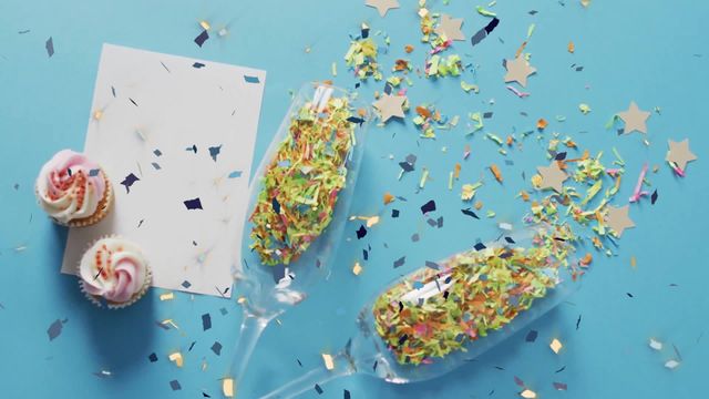 Bright festive arrangement features champagne glasses filled with multicolored confetti, accompanied by cupcakes, and a blank card on a vivid blue background. This lively concept can be used in promotional materials for parties or celebrations, digital greetings focusing on festive cheer, or invitations and announcements with a playful confetti theme.