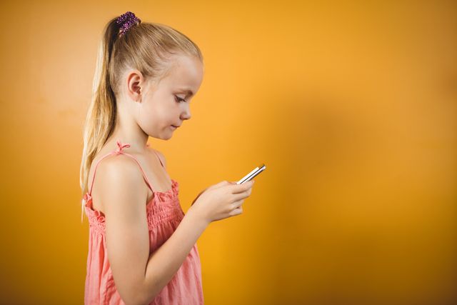 Young girl with a ponytail using a smartphone against an orange background. Ideal for illustrating concepts of children and technology, modern communication, and digital lifestyles. Suitable for educational materials, technology-related articles, and advertisements targeting young audiences.