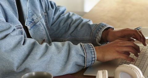 Mid-adult man typing on laptop at wooden table wearing denim jacket and headphones