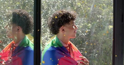 Young man wearing rainbow pride flag sitting by bus window with reflection, gold stars