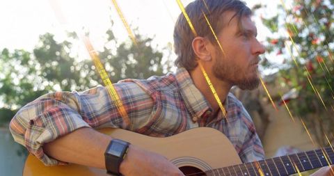 Playing acoustic guitar in sunlit backyard during golden hour, relaxed bearded man in plaid shirt