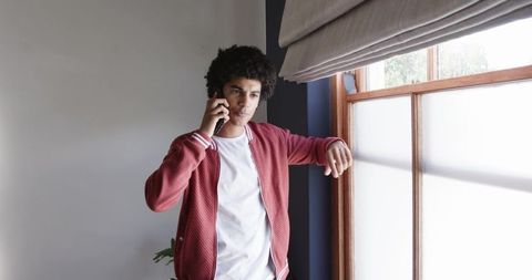 Young man talking on smartphone leaning on window sill wearing red bomber jacket