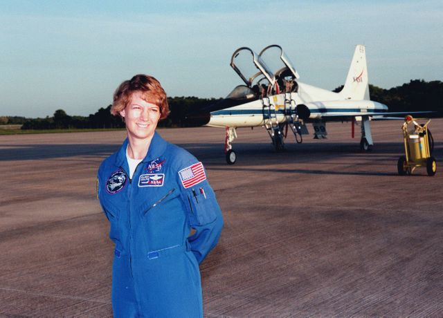 STS-93 Commander Eileen Collins poses for photographers in the early morning sun after landing at Kennedy Space Center's Shuttle Landing Facility (SLF) aboard a T-38 jet aircraft (background). She and other crew members Pilot Jeffrey S. Ashby and Mission Specialists Steven A. Hawley (Ph.D.), Catherine G. "Cady" Coleman (Ph.D.) and Michel Tognini of France, with the Centre National d'Etudes Spatiales (CNES), are arriving for pre-launch activities. Collins is the first woman to serve as mission commander. This is her third Shuttle flight. The primary mission of STS-93 is the release of the Chandra X-ray Observatory, which will allow scientists from around the world to study some of the most distant, powerful and dynamic objects in the universe. The new telescope is 20 to 50 times more sensitive than any previous X-ray telescope and is expected to unlock the secrets of supernovae, quasars and black holes