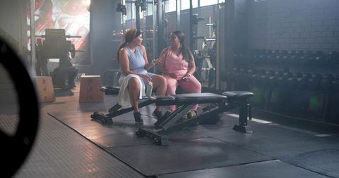 Diverse women chatting on gym bench during workout break, friendly fitness camaraderie