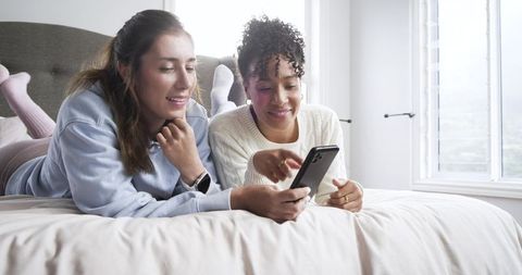 Two women lying on bed sharing smartphone, relaxing in bright minimal bedroom