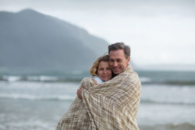 Romantic senior couple hugging while wrapped in a blanket on a beach. They are standing close to the ocean with mountains in the background, creating a serene and intimate moment. This image is perfect for use in advertisements for retirement communities, travel destinations, and lifestyle blogs that focus on love and relationships in later life.