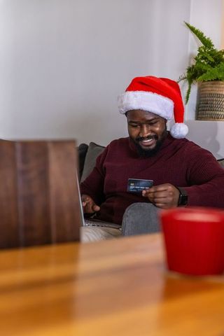 Black man in santa hat shopping online with credit card on sofa