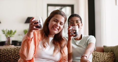 Two women smiling and clinking double-walled glass mugs while relaxing on cozy bobble sofa