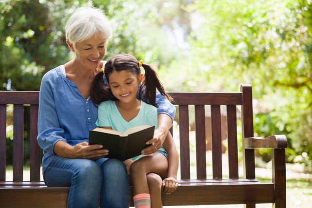 Elderly woman reading a book to her young granddaughter while sitting on a wooden bench in a garden. Ideal for use in family-oriented advertisements, educational materials, and content promoting intergenerational bonding and outdoor activities.