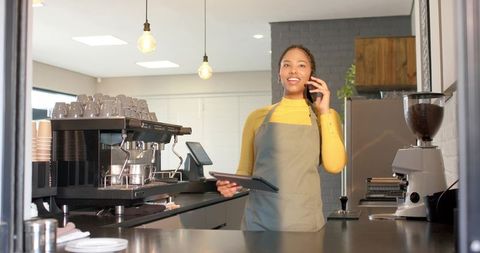 African american barista talking on phone while holding tablet behind modern cafe counter