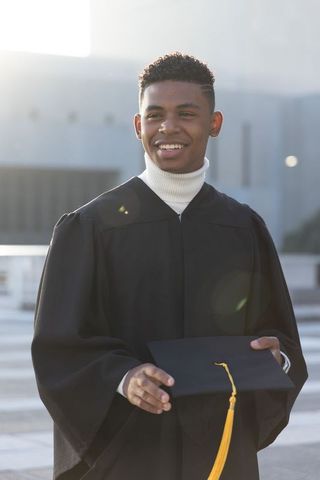 Smiling african american graduate holding mortarboard on sunlit modern campus plaza