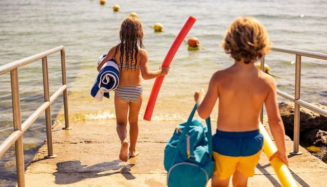 Children walking on pier toward sea carrying pool noodles, towel and beach duffel bag