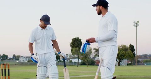 Diverse cricket teammates walking off grassy pitch at dusk holding bats and helmets