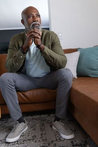 Mature african american man sitting on sofa gazing upward, hands clasped, contemplative