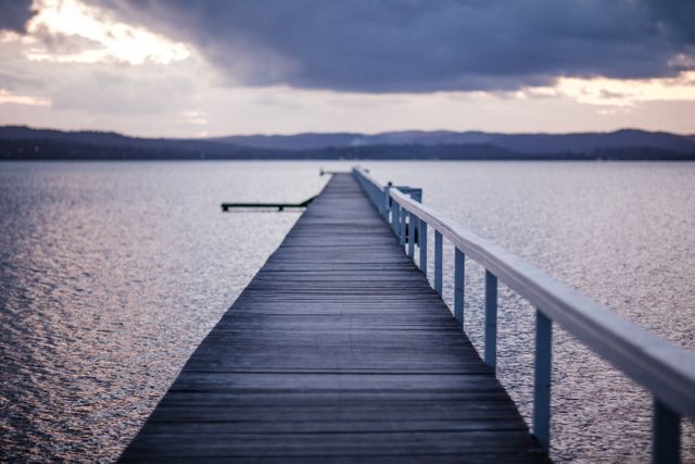 A long wooden pier extending into calm waters at sunset with mountains in the background and dusk clouds filling the sky. This image evokes serenity and solitude, and could be used for concepts related to relaxation, mindfulness, or travel. Ideal for backgrounds, screensavers, mindfulness blog posts, and relaxation videos.
