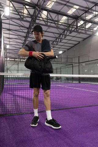 Male athlete standing at net on purple indoor tennis court holding duffel and towel wearing cap