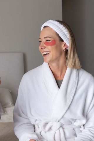 Woman enjoying at-home skincare routine wearing red under-eye patches and plush robe
