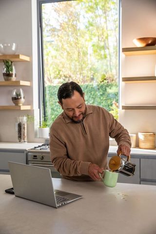 Mature asian man pouring coffee while working remotely in bright modern kitchen with laptop