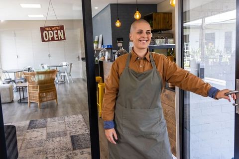Smiling barista opening cafe door welcoming customers at local coffee shop with open sign