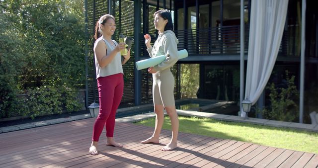 Two women in comfortable yoga attire standing on a sunlit wooden deck, enjoying smoothies and engaging in a friendly conversation. They appear to be discussing their exercise routine or planning the day ahead. The lush green surroundings evoke a serene and healthy environment, ideal for fitness and wellness activities. This image is suitable for advertising wellness programs, fitness products, or promoting a healthy lifestyle.