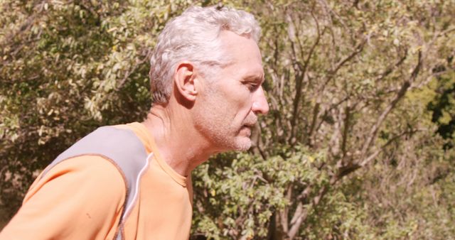 Senior man wearing an orange shirt, looking focused while engaging in an outdoor activity. He appears to be in a natural environment with dense vegetation and trees in the background. This image is ideal for promoting active lifestyles, wellness, or fitness among elderly individuals. It can be used in health-related articles, advertisements for outdoor activities or fitness programs for seniors, and general content focusing on healthy aging.