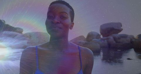 Smiling woman in blue swimsuit standing on coastal boulders at twilight with lens flare