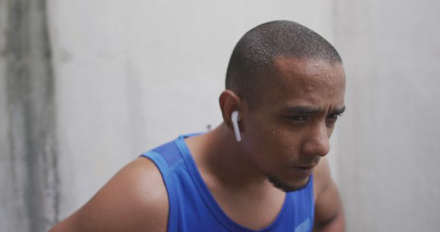 Young man wearing a blue tank top sweating during an intense workout, listening to music via wireless earbuds. Could be used for advertising athletic gear, promoting a healthy lifestyle, or illustrating the concept of determination and hard work.