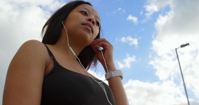 Woman enjoying music on earphones while standing outside on a sunny day with a blue sky and clouds. Perfect for themes related to relaxation, leisure, urban lifestyle, outdoor activities, and music enjoyment.