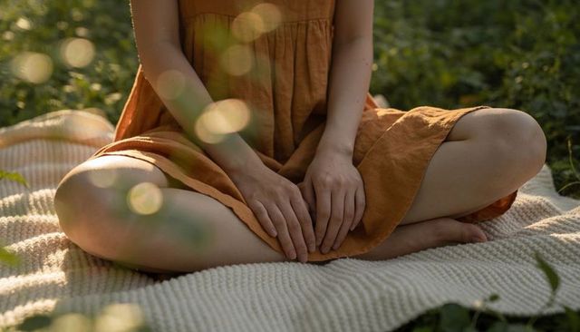 Woman sitting crosslegged in rust orange dress on cream blanket with sun bokeh