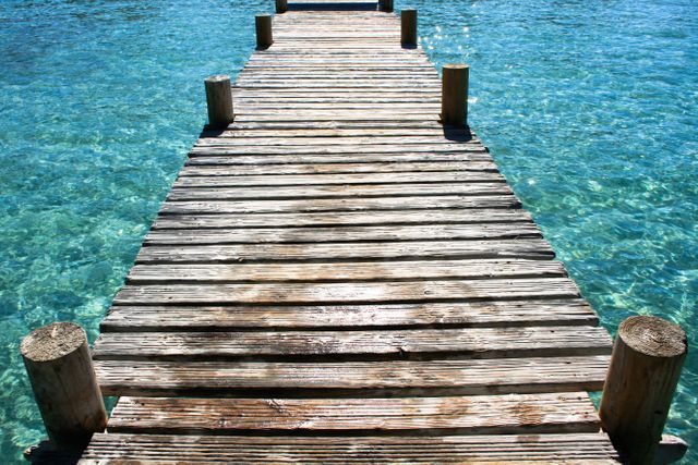 Wooden pier extends over crystal clear blue water, creating serene and inviting scene. Ideal for advertising tropical vacations, relaxation destinations, and beach adventures. Suitable for travel blogs, seaside resorts, and summer-themed promotions.