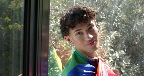 Young Man Draped in Rainbow Flag by Sunlit Window