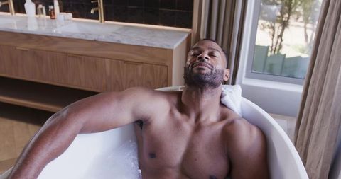 Mature man relaxing in freestanding bathtub with towel near marble vanity and window