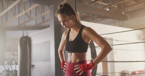 Focused female boxer pausing during intense boxing workout in gym ring with red hand wraps