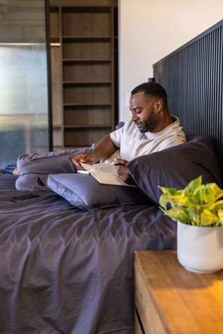 Mature african american man reclining on bed writing in notebook with smartphone, cozy bedroom