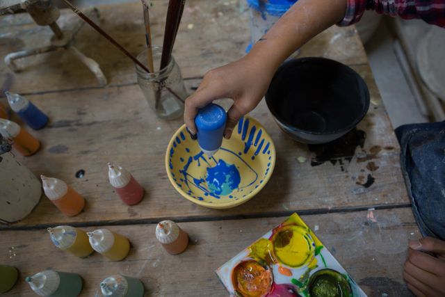 Young girl decorating a ceramic bowl with blue paint in a pottery workshop. Various paint bottles and brushes are scattered on the wooden table, showcasing a creative and artistic environment. Ideal for use in articles or advertisements about children's art activities, pottery classes, creative hobbies, or educational programs.