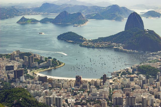 Aerial view of Rio de Janeiro with iconic Sugarloaf Mountain and Guanabara Bay in the background. High-rise buildings, verdant hills, and numerous boats in the bay create a picturesque scene. Suitable for travel brochures, tourism advertisements, geography projects, and presentations focusing on South American destinations.