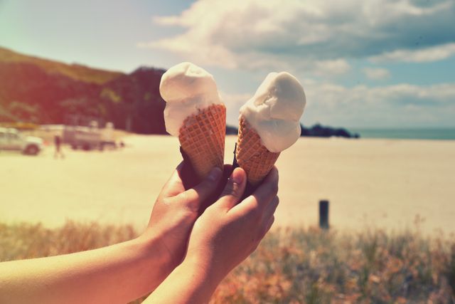Hands holding ice cream cones in waffle cones with sandy beach in background on a sunny day. Image capturing essence of summer, vacation and relaxation. Ideal for travel websites, ice cream advertisements, summer events promotions, food blogs, family vacation brochures, and leisure themed content.