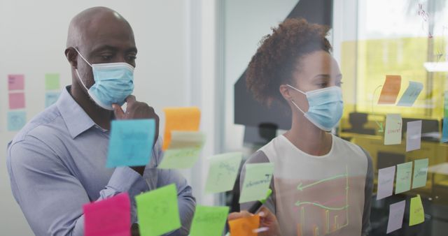 Diverse businessman and businesswoman in face masks brainstorming by board with memo notes in office. business professionals working in modern office during covid 19 coronavirus pandemic.
