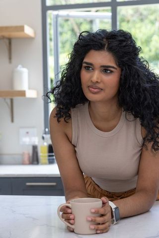 Indian woman leaning on marble counter holding mug wearing smartwatch in sunlit kitchen