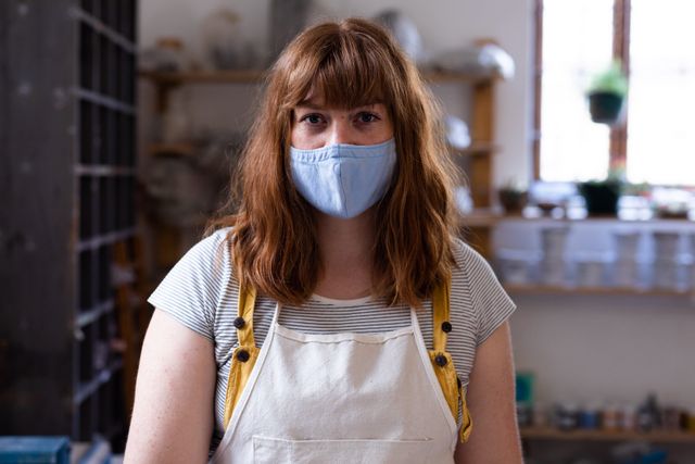 Portrait of caucasian female potter wearing face mask looking at camera. health and hygiene at pottery studio during coronavirus covid 19 pandemic.