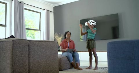 African american mother supervising daughter using vr headset while holding tablet in living room