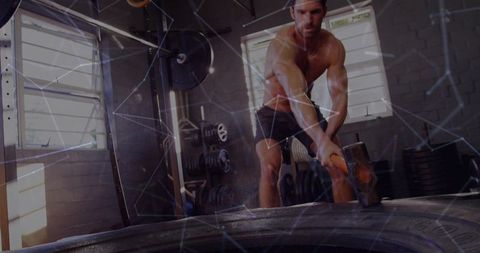 Shirtless man wearing black shorts smashing tractor tire with sledgehammer in garage gym
