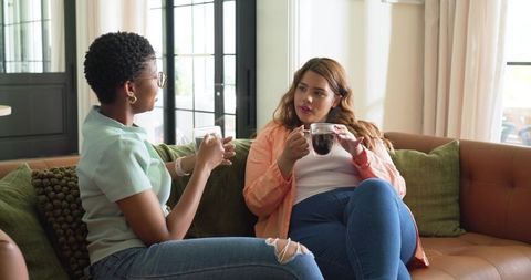 Diverse friends chatting and sipping coffee on leather couch in bright living room