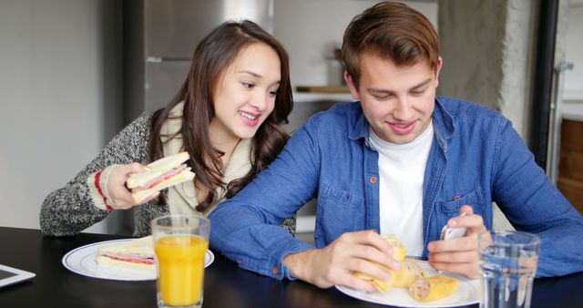Young couple sitting together at kitchen table, enjoying breakfast. Man checking phone while eating; woman looking over and holding sandwich, both smiling. Glass of orange juice on table. Ideal for lifestyle, relationship, and domestic scenes.