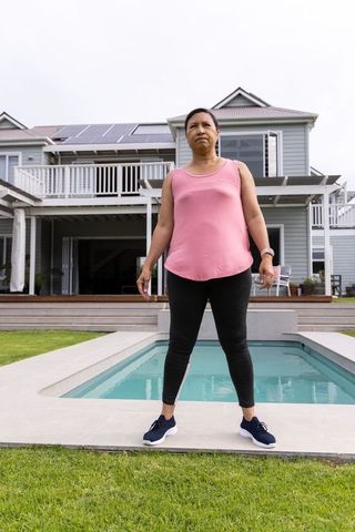 Mature woman standing at pool edge in modern backyard with deck, pergola and solar panels
