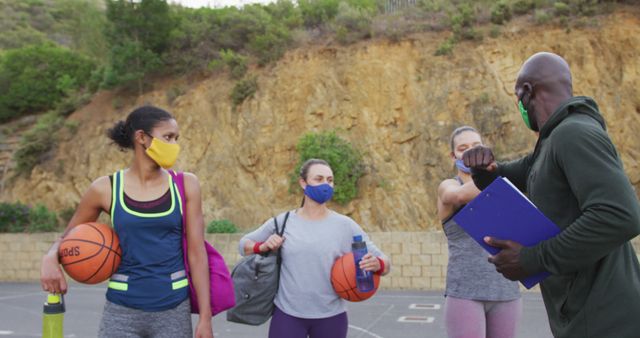 Diverse female basketball team and male coach wearing face masks greeting with elbows. basketball training at an outdoor urban court during coronavirus covid 19 pandemic.
