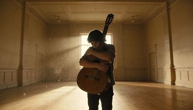 Teen embracing classical guitar in sunlit hall with dust particles and backlight