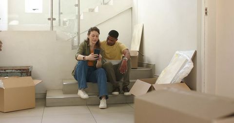 Young couple unpacking new home using smartphone on staircase surrounded by moving boxes