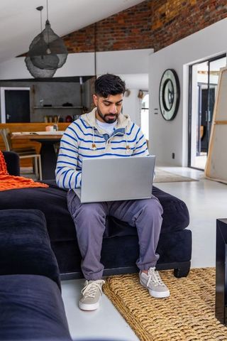 Indian man working on laptop in modern industrial loft home office on navy sofa